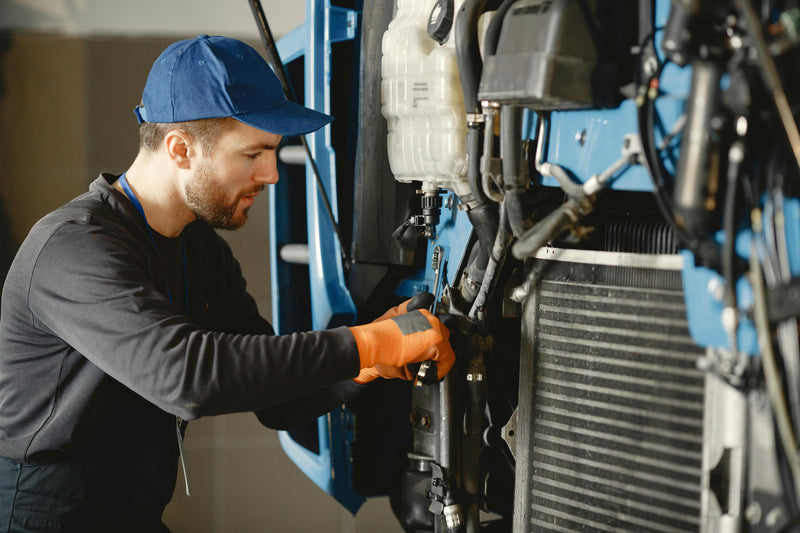 Mechanic working on a vehicle's engine with a blue cap and black jacket.